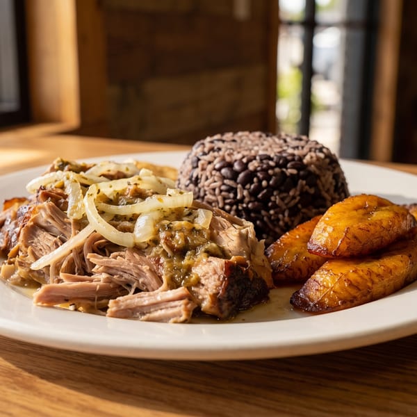 A plate of shredded pork with onions, rice and beans, and fried plantains on a wooden table.